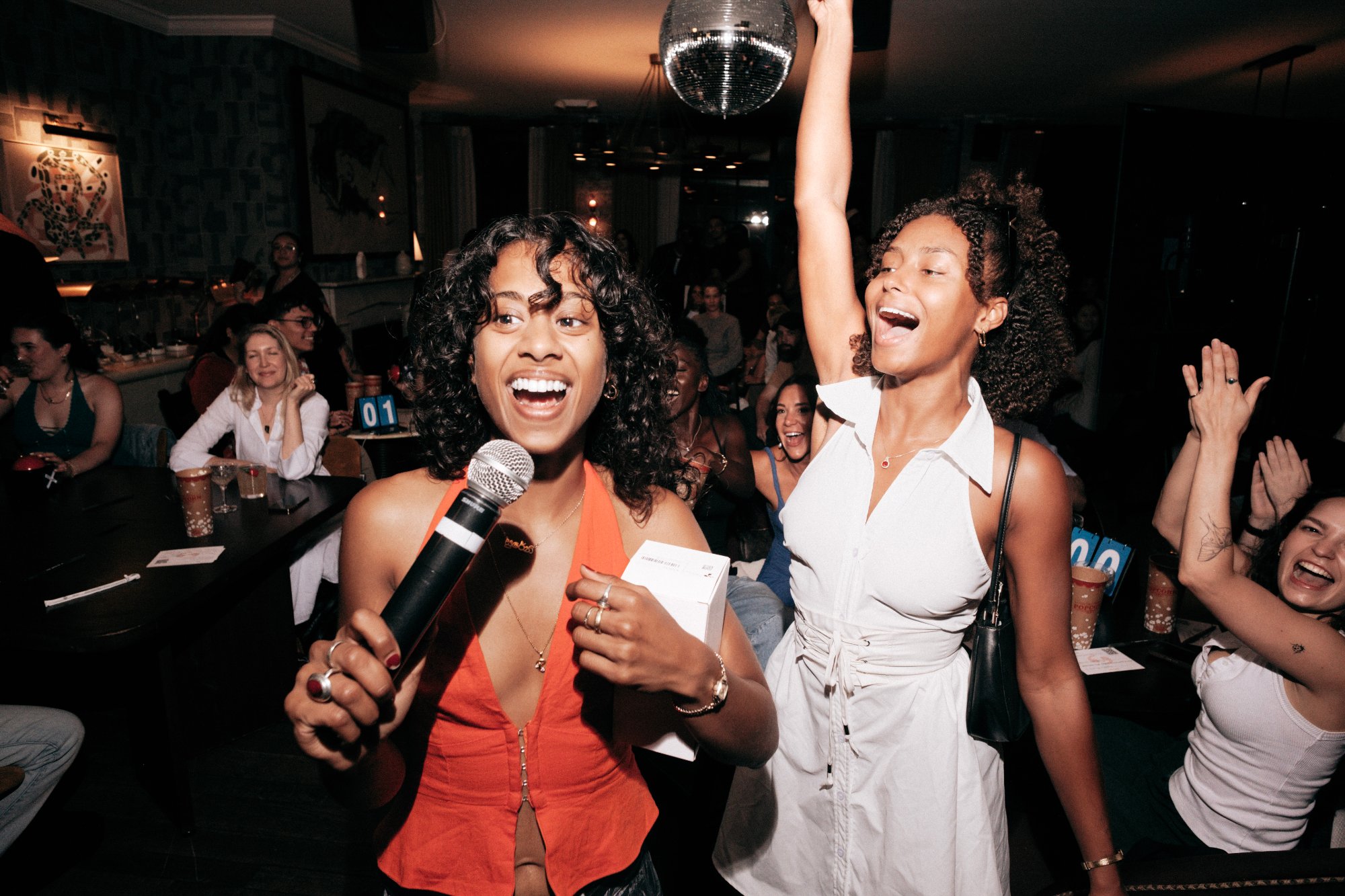 A performer in an orange halter top laughs mid-song with a mic in hand while a friend beside her throws an arm up cheering, in a warm, low-lit members' club room with a disco ball overhead and a crowd of seated guests behind.