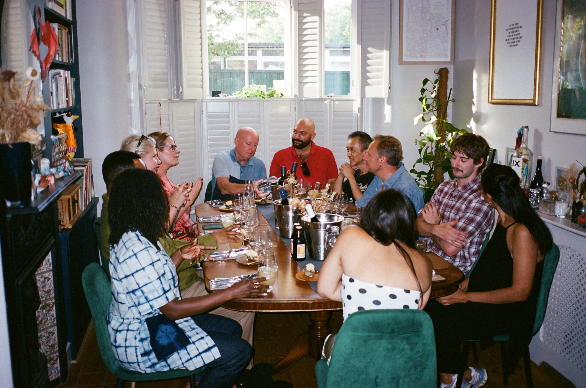 Guests around a long wooden dinner table in a sunlit living room, wine coolers and glasses down the middle, evening light through shuttered windows.